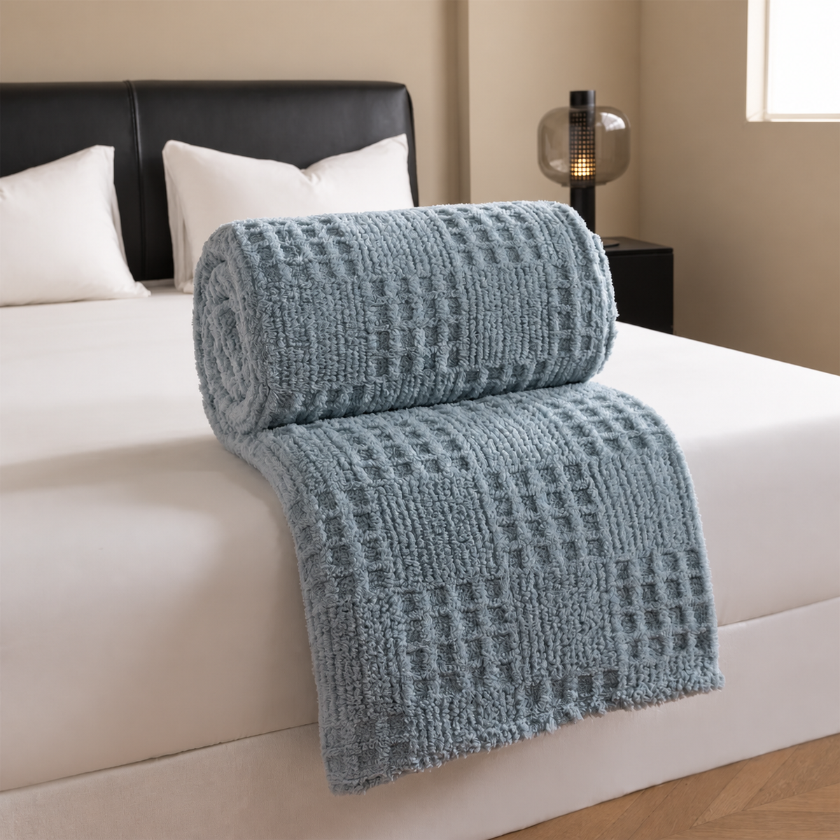 Plaid bleu gris jacquard gaufré roulé sur un lit blanc dans une chambre beige